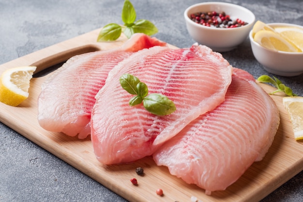 Filé de peixe cru de tilápia em uma tábua com limão e especiarias. Mesa escura