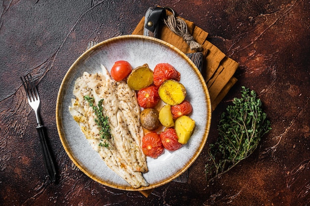 Filé de peixe Pollock assado no prato com guarnição de tomate e batata. Fundo escuro. Vista do topo.