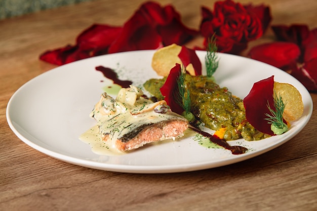 Filé de peixe vermelho delicioso com molho branco com legumes em molho verde em um prato branco. Um belo prato de restaurante decorado com pétalas de rosa.