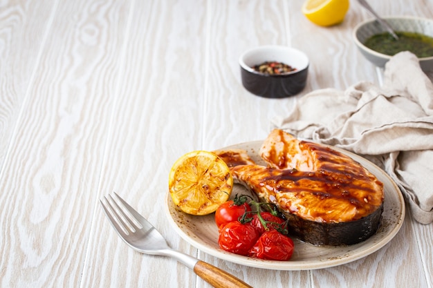 Filé de salmão grelhado com molho teriyaki, legumes e limão servido em um prato de cerâmica em uma mesa rústica de madeira branca em ângulo, foco seletivo com espaço para texto