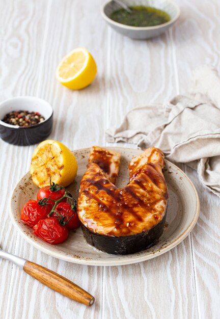 Filé de salmão grelhado com molho teriyaki, legumes e limão servido em um prato de cerâmica em uma mesa rústica de madeira branca em ângulo, foco seletivo