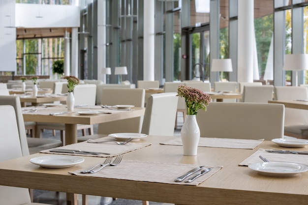 Fileira de mesas de jantar servidas com pratos, guardanapos e vasos com flores dentro de um luxuoso restaurante contemporâneo de grande centro de negócios