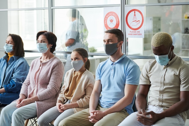 Foto fileira de pessoas de várias idades e etnias com máscaras protetoras esperando sua vez de receber a vacinação contra cobiça enquanto estão sentadas no hospital