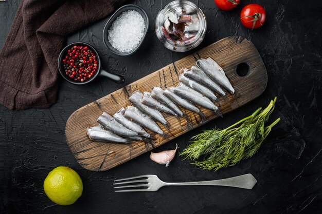 Filete de anchoas saladas en escabeche, sobre tabla de cortar de madera, sobre mesa negra con hierbas e ingredientes, vista superior plana