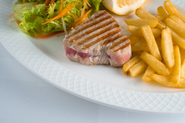 Filete de atún a la parrilla con ensalada de patatas fritas de zanahorias, chirivías, lechuga y limón en un plato blanco sobre fondo blanco.