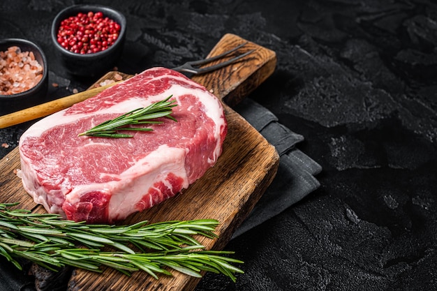 Filete de chuletón crudo carne de vacuno marmolada sobre tabla de madera con romero Fondo negro Vista superior Espacio de copia