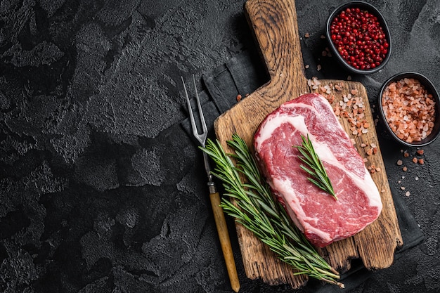 Filete de chuletón crudo carne de vacuno marmolada sobre tabla de madera con romero Fondo negro Vista superior Espacio de copia