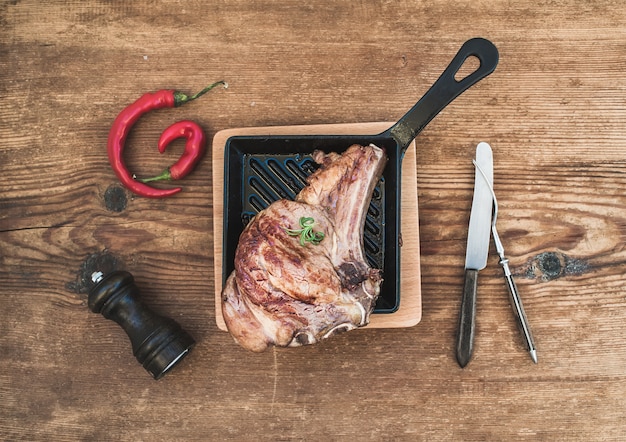 Filete de costilla de carne cocida con especias, chile rojo, romero y cubiertos vintage en sartén sobre mesa de madera rústica, vista superior.