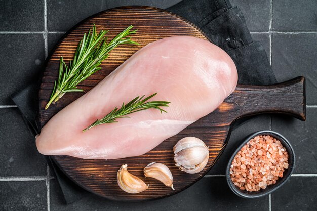 Filete de pecho de pollo crudo en una tabla de madera con romero y ajo carne de aves de corral de fondo negro Vista superior