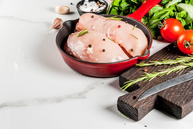 Filete de pechuga de pollo fresco crudo con hierbas y especias para cocinar, en sartén de hierro fundido, mesa de mármol blanco, espacio de copia