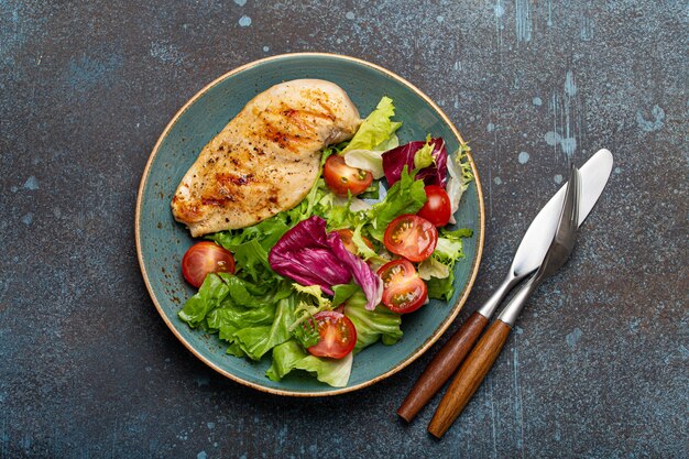 Filete de pechuga de pollo a la parrilla con ensalada de verduras de tomates frescos y hojas de ensalada en un plato azul sobre fondo de piedra rústica desde arriba, cena saludable o comida dietética