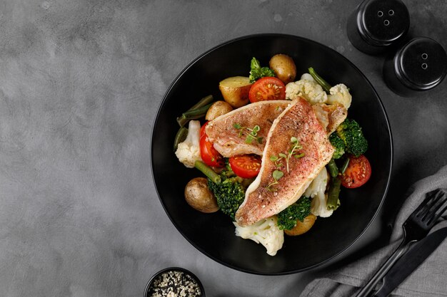 Filete de perca de pescado con verduras brócoli coliflor tomates patatas en un plato oscuro