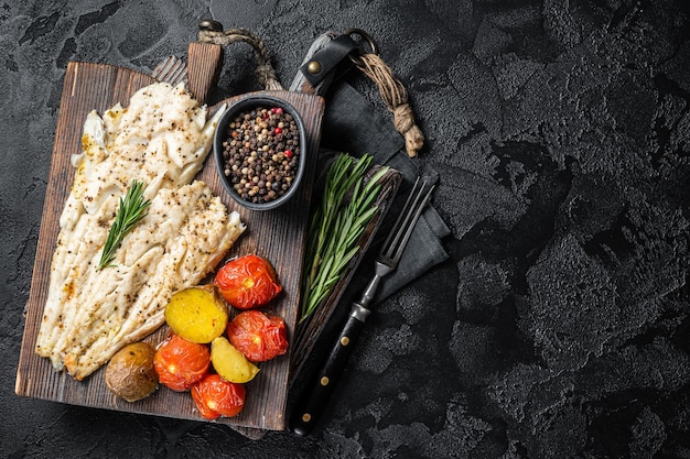 Filete de pescado de eglefino al horno sobre tabla de madera con tomate y patata Fondo negro Vista superior Espacio de copia