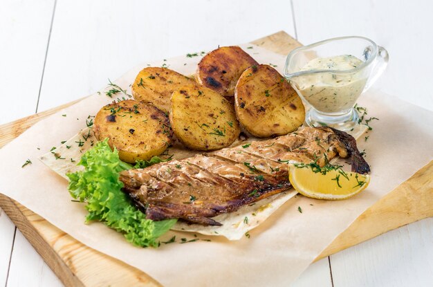 Filete de pescado a la plancha con patatas al horno con salsa sobre una tabla de madera. Fondo de madera blanca