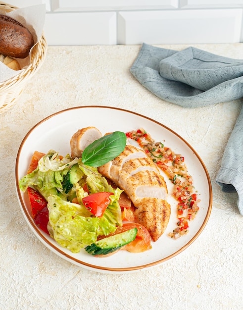 Filete de pollo a la plancha con verduras y salsa bearnesa. En un plato, junto a él, hay pan y una servilleta azul. Fondo claro.