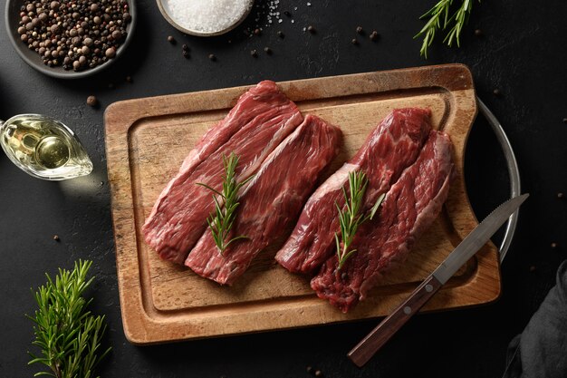 Filete de ribeye crudo en tabla de cortar con romero sobre fondo negro.