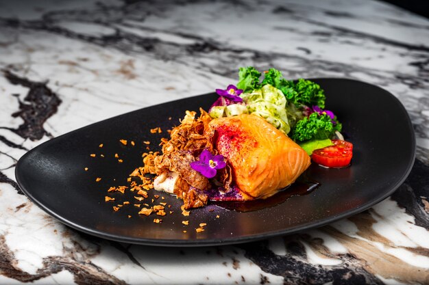 Filete de salmón al horno con verduras y guarnición de flores servido en una placa negra