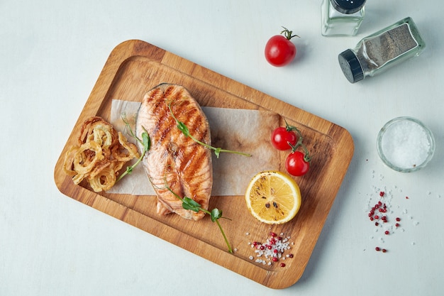 Filete de salmón a la plancha con verduras y cebollas fritas en una tabla de madera sobre una mesa blanca. Vista superior