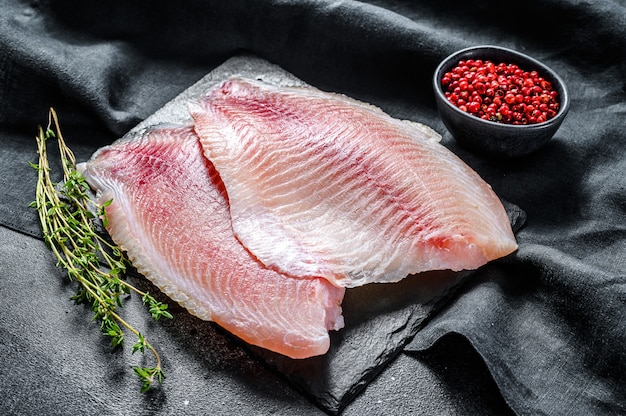 Filete de telapia crudo en una tabla de cortar con tomillo y pimienta rosa. fondo negro. vista superior