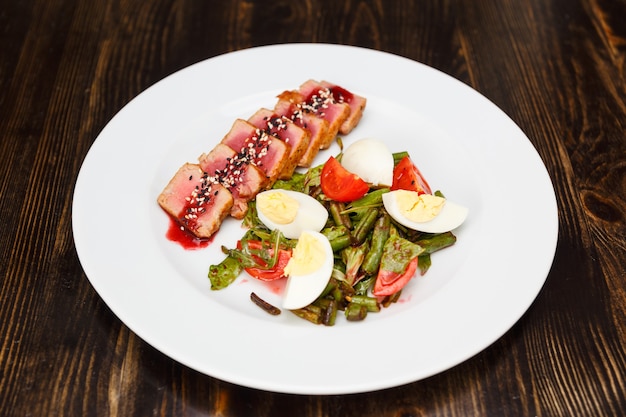 Filete de ternera medio trasero cortado en rodajas y rociado con salsa de sésamo en un plato blanco. Adorne con espárragos cocidos, huevos y tomates.