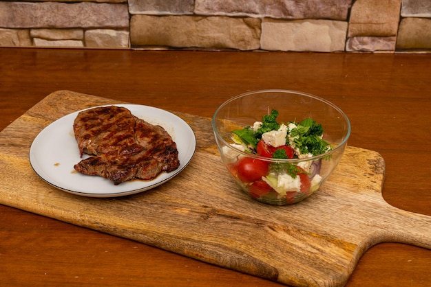 Filete de ternera a la parrilla en un plato y una taza de ensalada en una tabla de cortar de madera