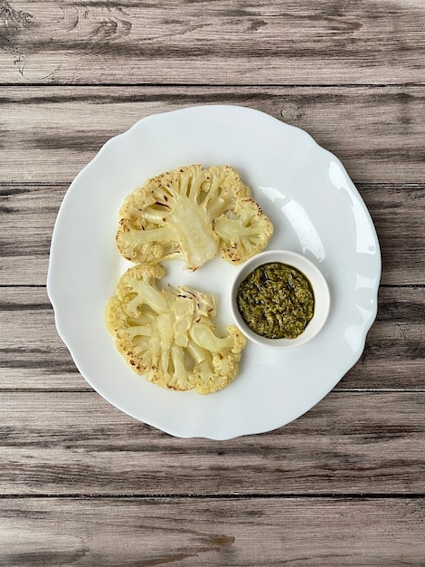 Filetes de coliflor con salsa de pesto en un plato blanco en una disposición vertical de la vista superior de la placa de madera