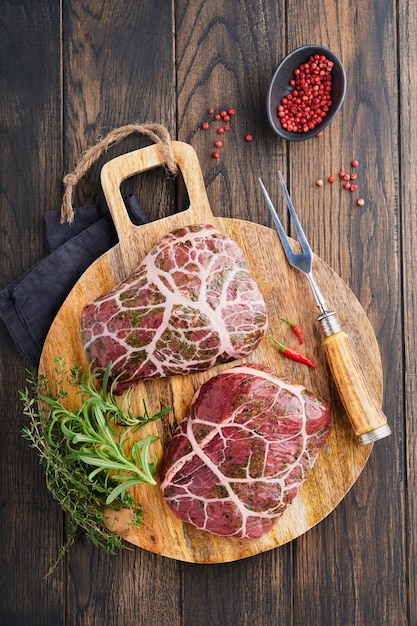 Filetes crudos bistec de vaquero crudo carne de res veteada con especias romero y pimienta en tablero de madera negra sobre fondo de mesa oscura de madera vieja Vista superior maqueta