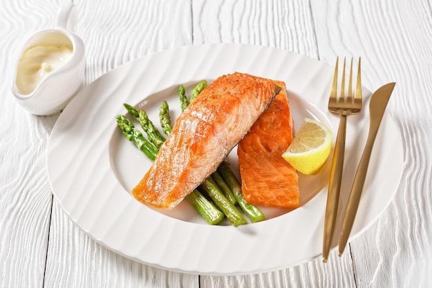 Filetes de salmão assado com aspargos grelhados e fatias de limão em um prato branco sobre uma mesa texturizada de madeira branca vista horizontal de cima