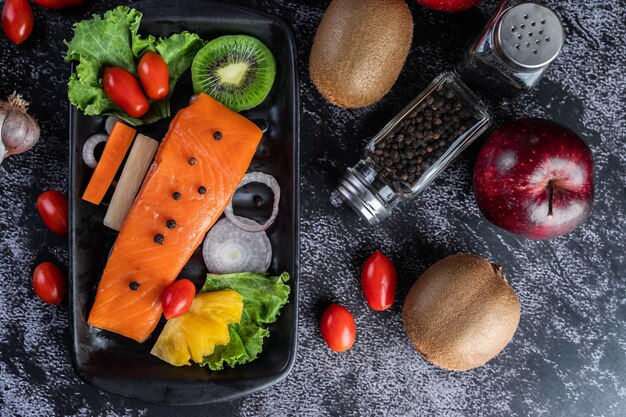 Foto filetes de salmão cru, pimenta, kiwi, abacaxi e romário em um prato e chão de cimento preto