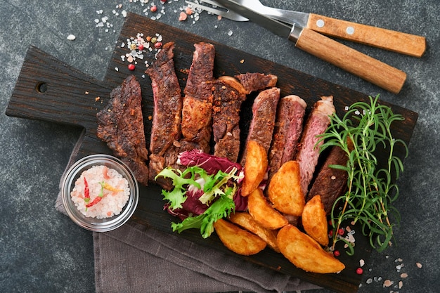 Filetes Filete de carne a la parrilla en rodajas New York Ribeye o Chuck roll con guarnición de ensalada y papas fritas en tablero de mármol negro sobre fondo de madera vieja Vista superior Maqueta