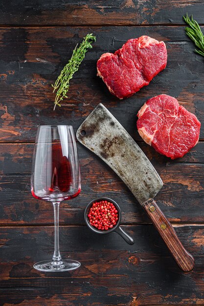 Filetes de grupa crudos con cuchillo de carnicero americano y dos vasos de vino tinto sobre fondo de madera vieja oscura, vista superior.