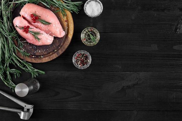 Filetes de pechuga de pollo cruda sobre tabla de cortar de madera con hierbas y especias sobre un fondo de madera negra. Vista superior con espacio de copia. Naturaleza muerta. Endecha plana