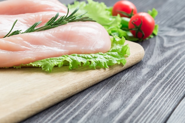 Filetes de pechuga de pollo cruda sobre una tabla para cortar. Taza de aceite con tomates