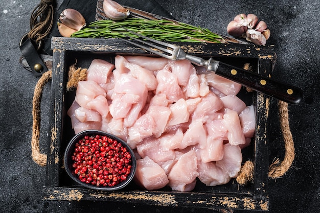 Filetes de pechuga de pollo en cubitos crudos en rodajas de carne de ave en bandeja de madera Fondo negro Vista superior