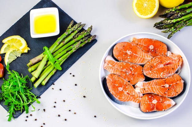 Filetes de salmón crudo fresco con verduras y especias: espárragos, tomates, granos de pimienta, rúcula, limón y aceite de oliva en la mesa gris. Concepto de dieta de alimentos saludables.