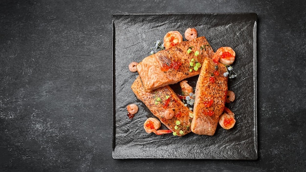 Filetes de salmón frito con camarones y caviar rojo en un plato negro sobre un fondo oscuro con espacio para texto