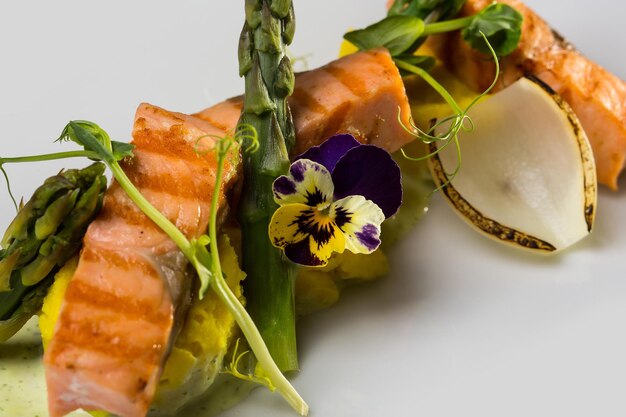 Filetes de salmón a la parrilla adornados con puré de salsa, espárragos de flores y verduras sobre fondo blanco.