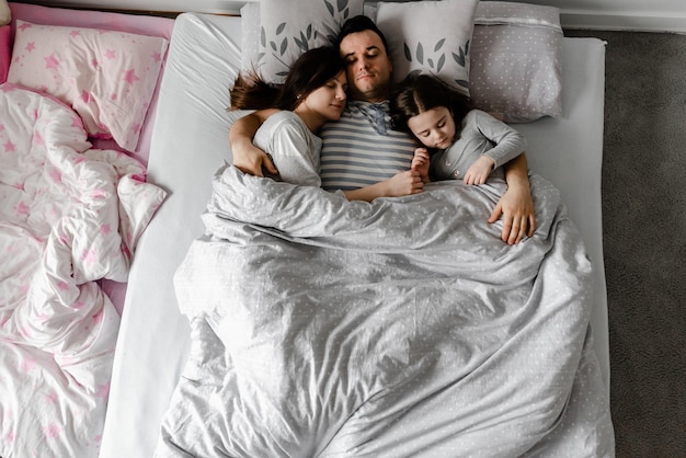 Filha com os pais na cama, o pai beija suavemente a esposa e a filha, fortes abraços familiares, fim de semana familiar na cama, família dormindo, filha dorme com pais jovens na cama