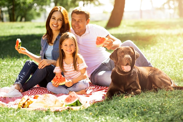 Filha com pais e cachorro comendo melancia no parque e olhando para a câmera