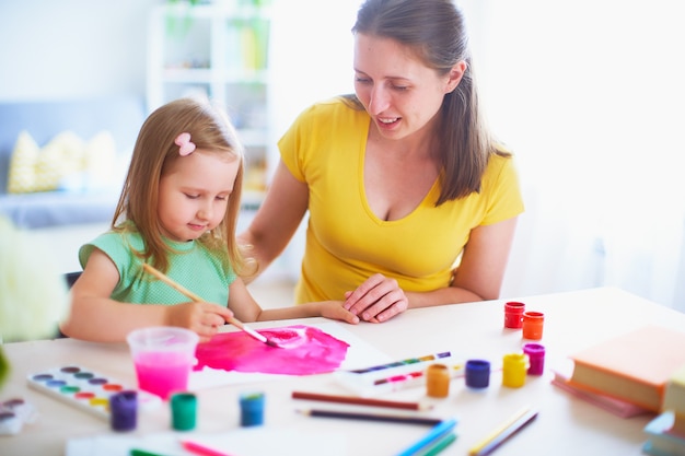 Filha de mãe pinta aquarela em uma folha de papel, sentado em casa à mesa em uma sala iluminada.
