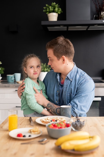 Filha exigente sentada nos joelhos do pai fazendo careta enquanto se recusa a comer o café da manhã feito pelo pai