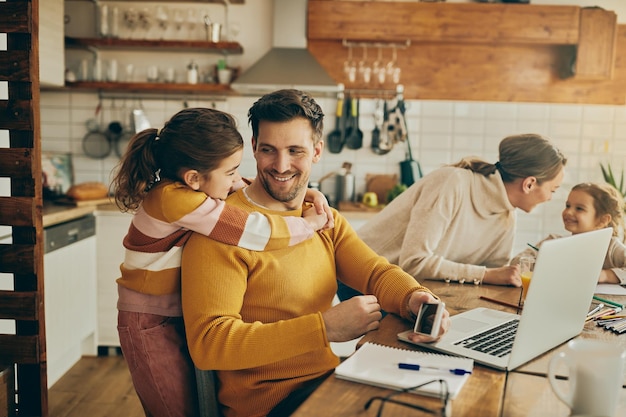 Filha feliz abraçando seu pai que está trabalhando no laptop em casa
