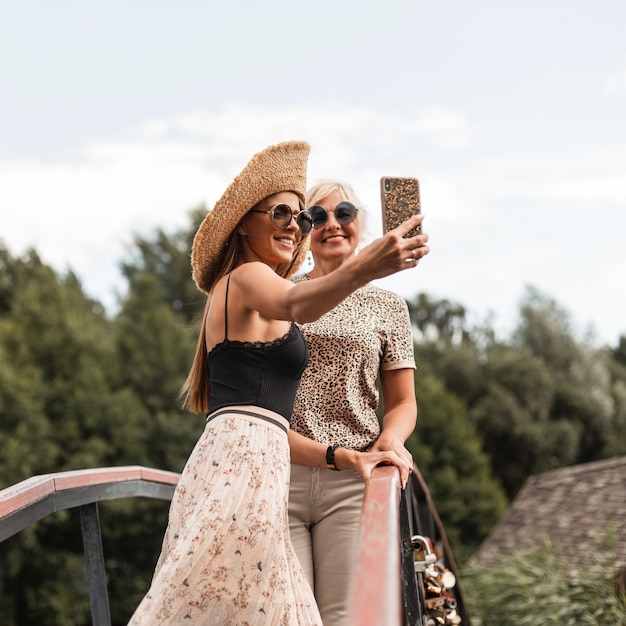 Filha feliz com a mãe sorridente em roupa da moda de verão com chapéu de palha e óculos escuros faz uma foto conjunta no smartphone e descansa na natureza do campo