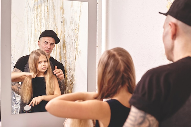 Filha fofa e seu pai tatuado estão brincando juntos perto de um espelho que o pai está fazendo com sua filha ...