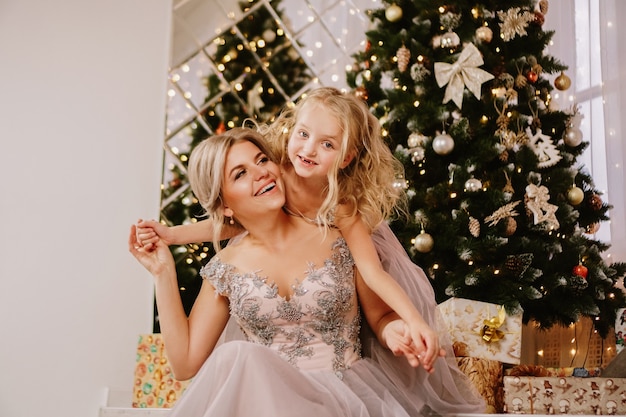Filha sorridente abraçando a mãe enquanto está sentada na árvore de Natal com caixas de presente