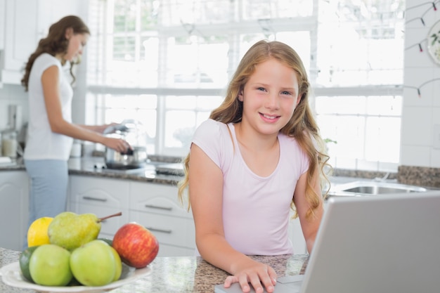 Filha, usando computador portátil, com, mãe, cozinhando alimento, em, fundo, em, cozinha