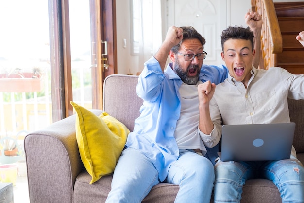 Filho adulto feliz e pai maduro dos anos 50 usando laptop comemorando o sucesso Duas gerações familiares de fãs de esportes assistindo jogo online ou jogo no computador fazendo gesto de vitória sim se divertindo