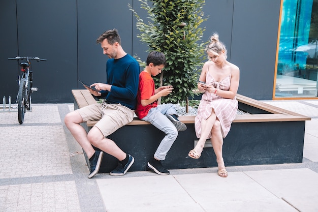 Filho e pais jovens usam tecnologia sem fio enquanto relaxam na praça da cidade num dia de verão. Foto de alta qualidade. Estilo de vida