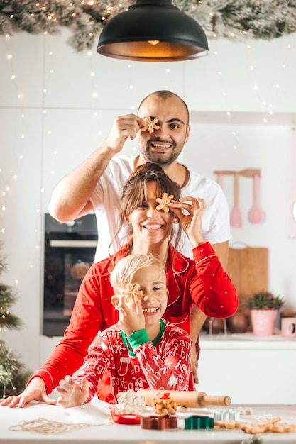 Filho, mãe e pai assam bolos de natal na cozinha sorrindo, olhando para a câmera de pijama