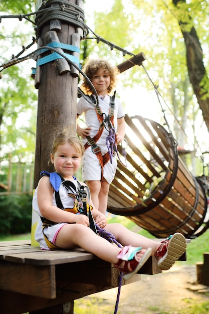 Filhos bonitos - um menino e uma menina em um parque de aventura estão envolvidos em escaladas ou ultrapassam obstáculos na estrada de corda.